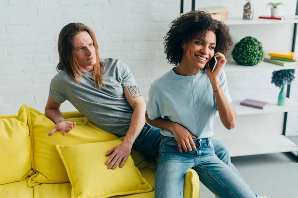 Upset young man gesturing by hand to smiling african american girlfriend talking on smartphone — Stock Photo