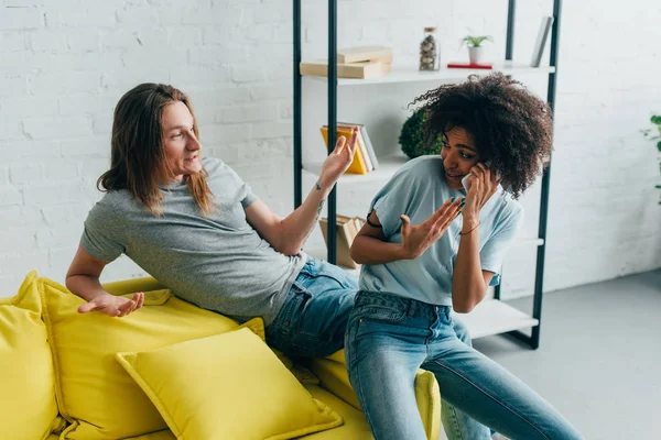 Smiling man with wide arms and african american girlfriend gesturing and talking on smartphone — Stock Photo