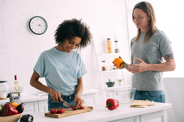 Femme afro-américaine souriante coupant des tomates cerises à bord tandis que petit ami debout près avec poivron à la main — Photo de stock