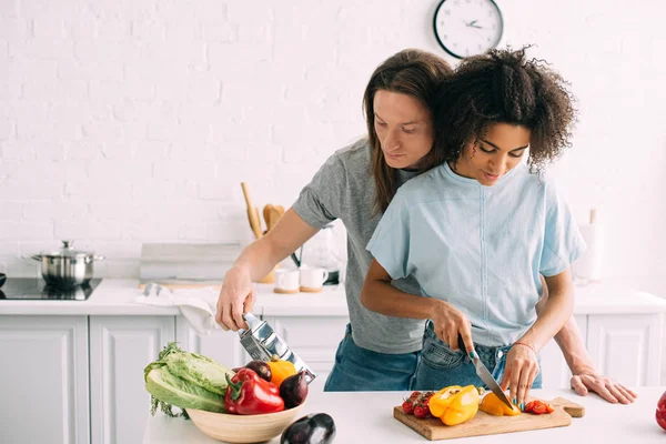 Jovem mulher cortando pimentão e namorado olhando ralador na cozinha — Fotografia de Stock