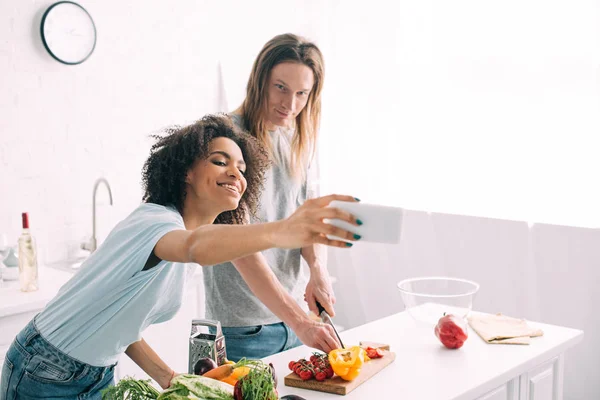Щаслива афроамериканка бере селфі на смартфон з хлопцем, який ріже овочі на кухні — стокове фото