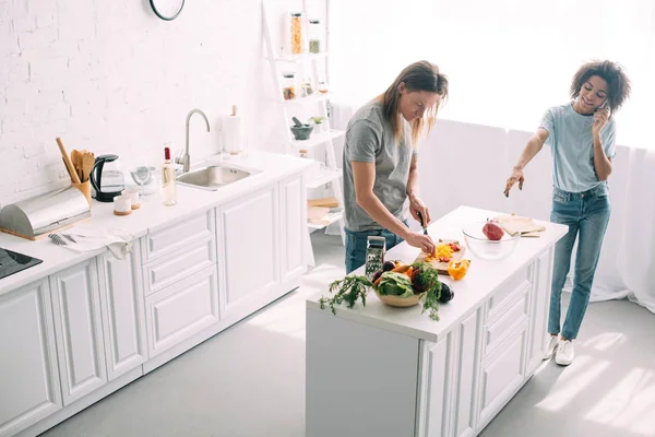 High angle view of young woman talking on smartphone and pointing by hand to boyfriend while he cutting vegetables at kitchen — Stock Photo