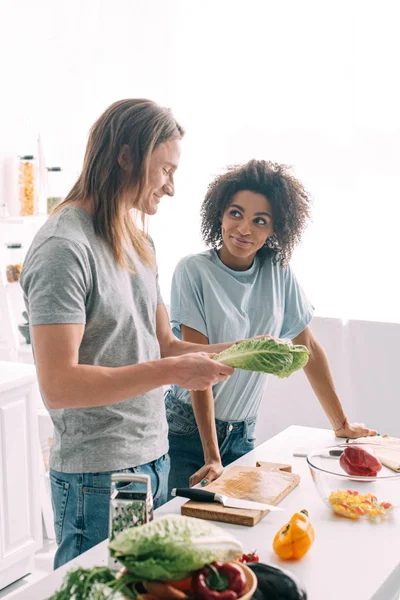 Jovem casal multiétnico de pé na cozinha com folhas de salada — Fotografia de Stock