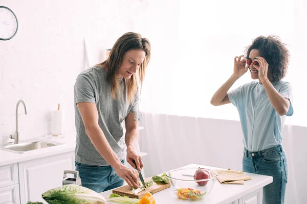 Heureuse femme afro-américaine couvrant les yeux par des tomates cerises et petit ami couper des feuilles de salade à la cuisine — Photo de stock