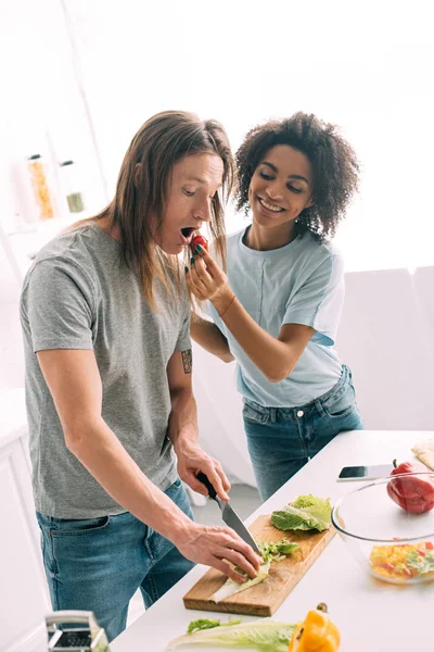 Sorridente donna afro-americana che nutre fidanzato mentre cucina in cucina — Foto stock