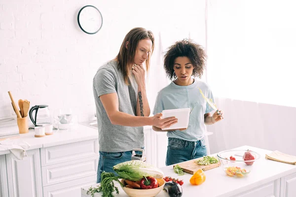 Jeune couple multiculturel regardant la recette sur tablette numérique et la cuisine à la cuisine — Photo de stock