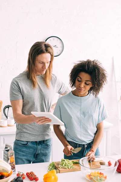 Man showing recipe on digital tablet to african american girlfriend while she cutting salad at kitchen — Stock Photo