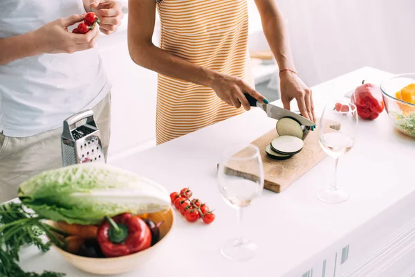 Cropped shot of woman cutting aubergine and boyfriend standing near with cherry tomatoes at kitchen — Stock Photo