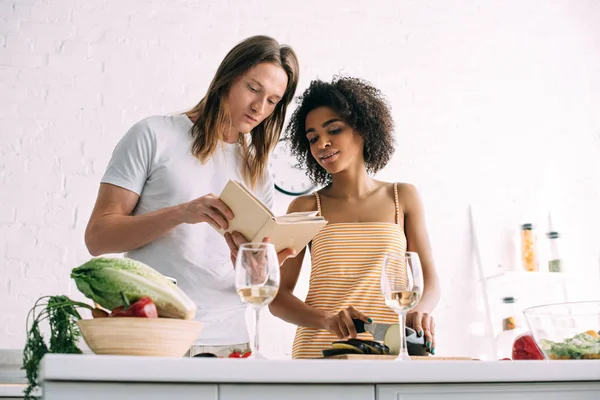 Joven pareja multicultural mirando receta en libro en la cocina - foto de stock