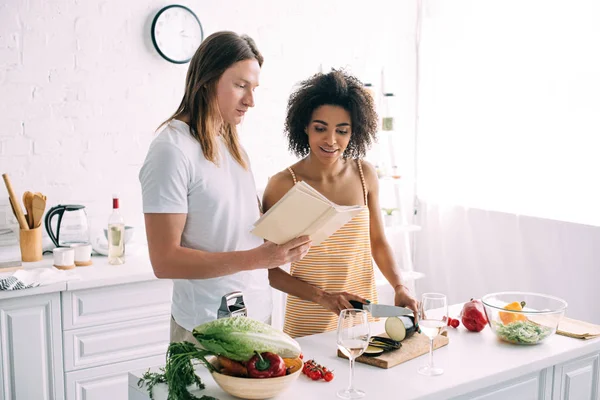 African american woman cutting aubergine and boyfriend showing recipe to her — Stock Photo