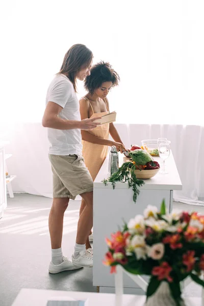 Side view of multicultural couple with recipe in book cooking at kitchen — Stock Photo