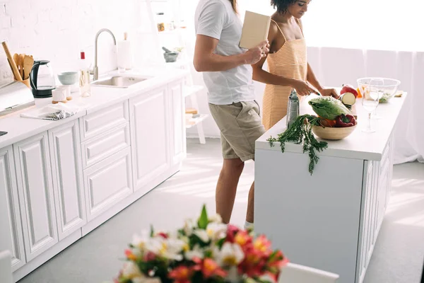Immagine ritagliata di uomo con ricettario e fidanzata che taglia verdure in cucina — Foto stock