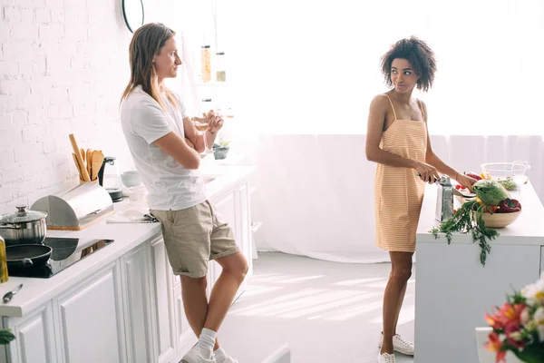 Joven con copa de vino y hablando con la novia afroamericana mientras ella corta verduras en la cocina - foto de stock