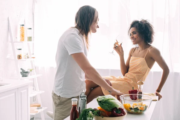 Smiling african american woman with wine glass sitting on table and talking to boyfriend at kitchen — Stock Photo