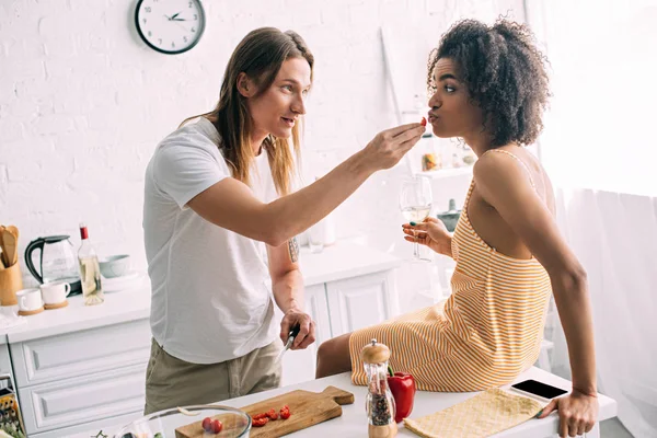 Young man feeding smiling african american girlfriend by cherry tomato at kitchen — Stock Photo