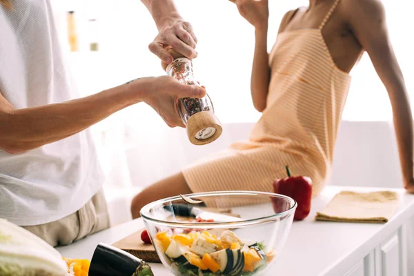 Cropped shot of man using pepper grinder over bowl with salad and woman sitting on table at kitchen — Stock Photo