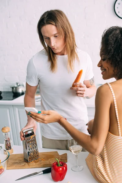 Sonriente mujer afroamericana mostrando receta en smartphone a novio con zanahoria y rallador en la cocina - foto de stock