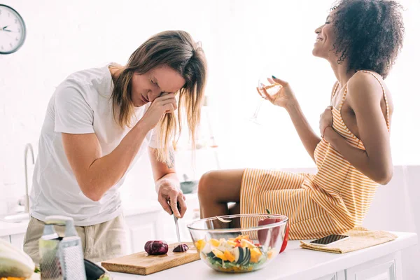 Femme afro-américaine souriante avec verre de vin assis sur la cuisine tandis que son petit ami coupant l'oignon et pleurant à la cuisine — Photo de stock