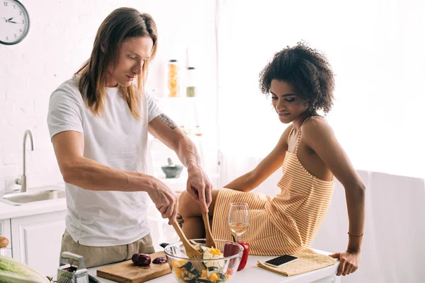 Африканская американская молодая женщина смотрит на парня, пока он готовит салат на кухне — стоковое фото