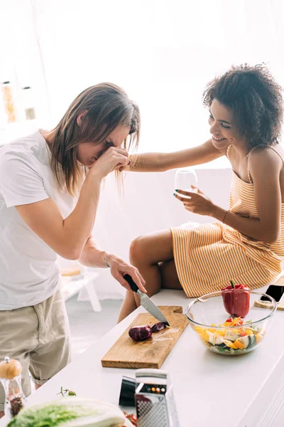 Sorridente afroamericano che tiene il bicchiere di vino e rallegra il fidanzato piangendo mentre taglia la cipolla in cucina — Foto stock