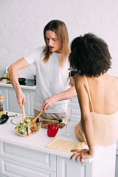 Vue arrière de la femme tirant petit ami sur smartphone pendant qu'il cuisine la salade à la cuisine — Photo de stock