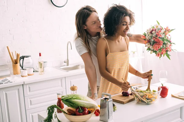Young man gifting bouquet of flowers to african american girlfriend while she cooking at kitchen — Stock Photo