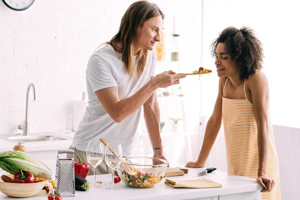 Young man feeding african american girlfriend by salad at kitchen — Stock Photo