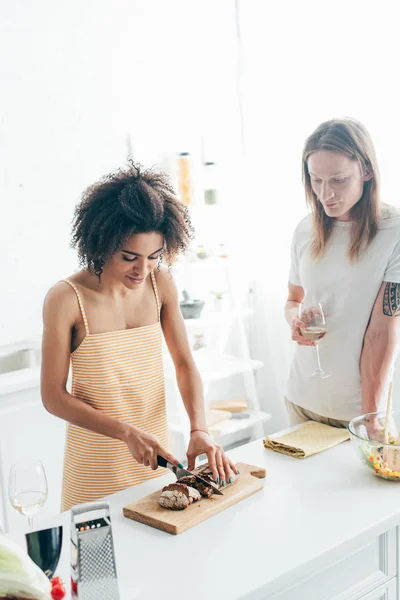 African american woman cutting bread and boyfriend standing near with wine glass — Stock Photo