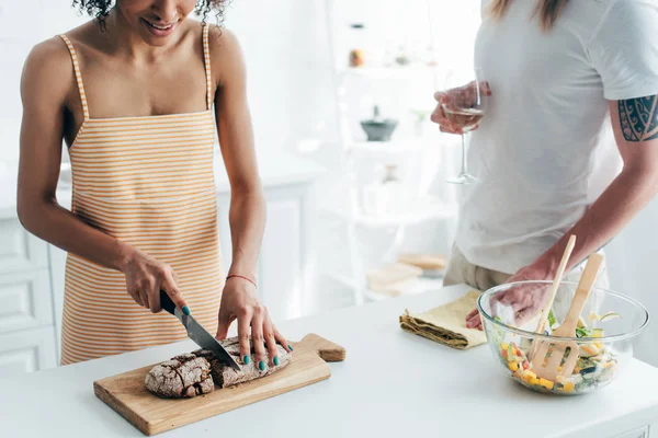 Imagem cortada de mulher cortando pão e namorado de pé perto com copo de vinho — Fotografia de Stock