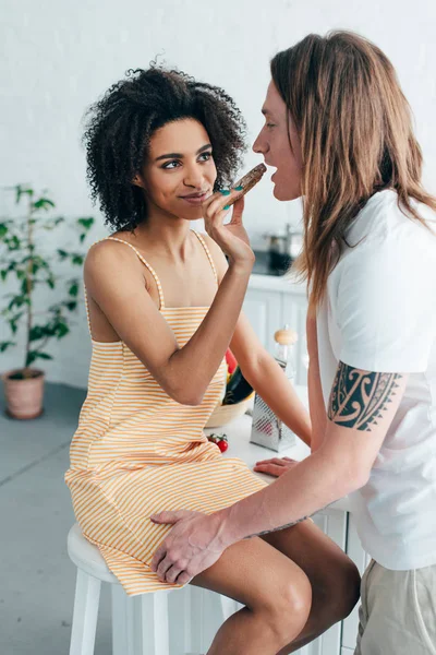 Sonriente africano americano mujer alimentación novio con tatuado mano en cocina - foto de stock