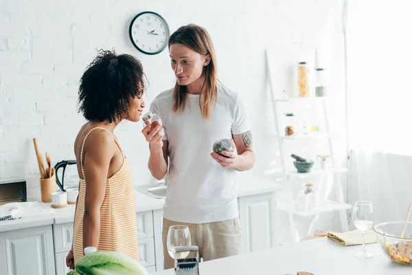 Young man with tattooed hand giving girlfriend to sniff spices at kitchen — Stock Photo