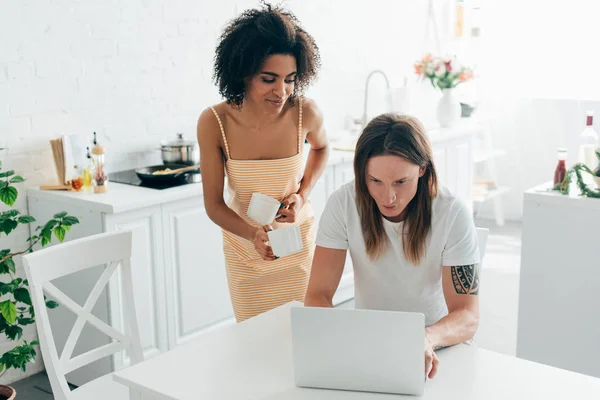 African american woman with coffee cups and boyfriend using laptop at kitchen — Stock Photo