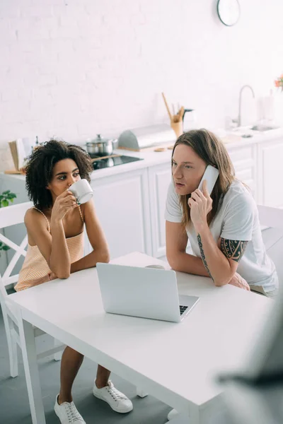 African american woman drinking coffee and boyfriend talking on smartphone at table with laptop — Stock Photo