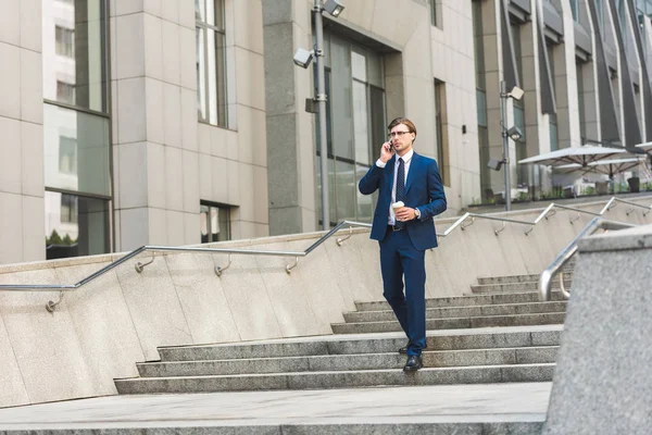 Businessman with coffee to go talking by phone while walking by stairs near business building — Stock Photo
