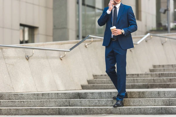 Cropped shot of businessman in stylish suit with coffee to go talking by phone while walking by stairs near business building — Stock Photo