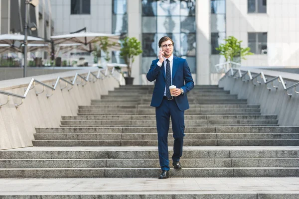 Sonriente joven hombre de negocios en traje elegante con café para ir a hablar por teléfono mientras camina por las escaleras cerca del edificio de negocios - foto de stock