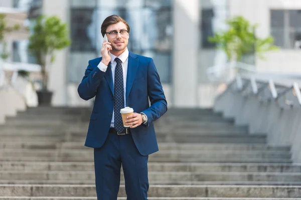 Sonriente joven hombre de negocios en traje elegante con café para ir a hablar por teléfono en las escaleras cerca del edificio de negocios - foto de stock