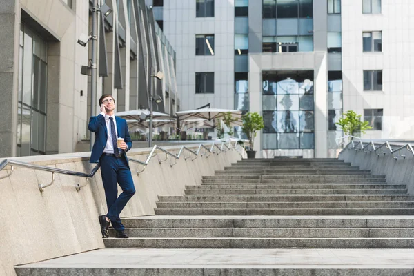 Handsome young businessman in stylish suit with coffee to go talking by phone on stairs near business building — Stock Photo