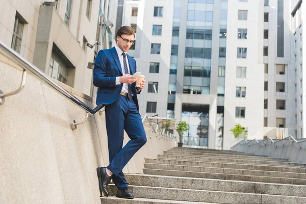 Attractive young businessman in stylish suit with paper cup of coffee using smartphone on stairs near business building — Stock Photo