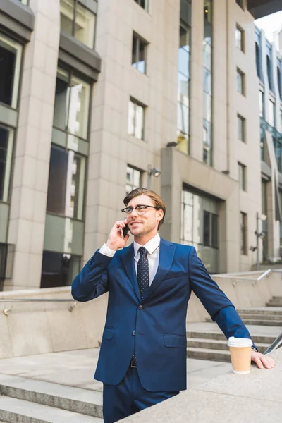 Giovane uomo d'affari sorridente con tazza di caffè di carta che parla per telefono e distoglie lo sguardo vicino all'edificio di affari — Foto stock