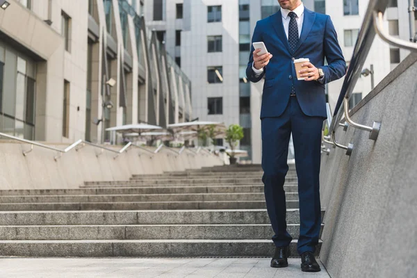 Обрізаний знімок бізнесмена в стильному костюмі з кавою, щоб піти за допомогою смартфона під час сходу сходами — стокове фото
