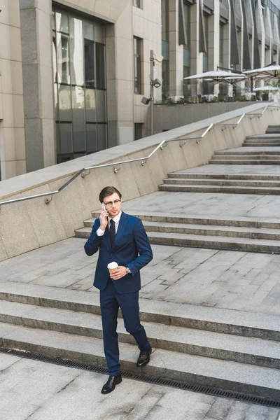 Joven hombre de negocios guapo en traje elegante con café para ir a hablar por teléfono mientras baja escaleras - foto de stock