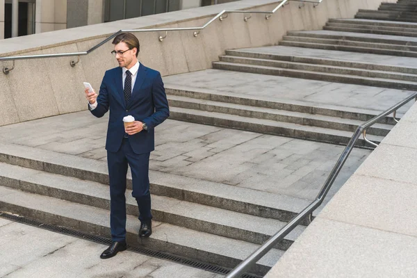 Sonriente joven hombre de negocios en traje elegante con café para ir usando el teléfono inteligente mientras baja las escaleras - foto de stock