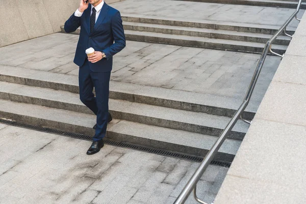 Recortado disparo de hombre de negocios en traje elegante con café para ir hablando por teléfono mientras baja escaleras - foto de stock