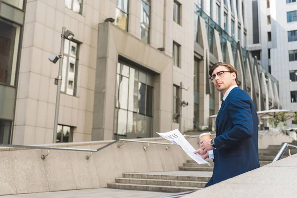 Joven hombre de negocios guapo con café para ir y periódico en el distrito de negocios - foto de stock