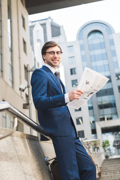Vista inferior de sonriente joven hombre de negocios en traje elegante con periódico en el distrito de negocios - foto de stock