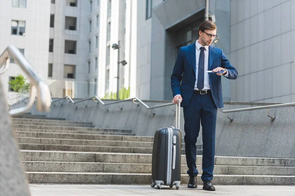 Bonito jovem empresário com bagagem e bilhetes de avião andando por distrito de negócios e olhando para assistir — Fotografia de Stock