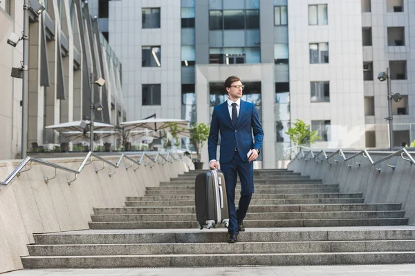 Joven hombre de negocios en traje elegante con equipaje y billetes de avión que baja escaleras en el distrito de negocios - foto de stock
