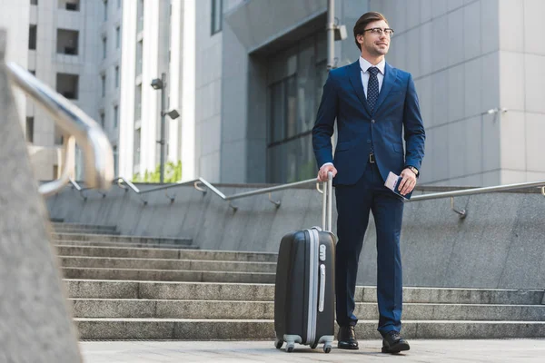 Successful businessman in stylish suit with luggage and flight tickets going down stairs in business district — Stock Photo