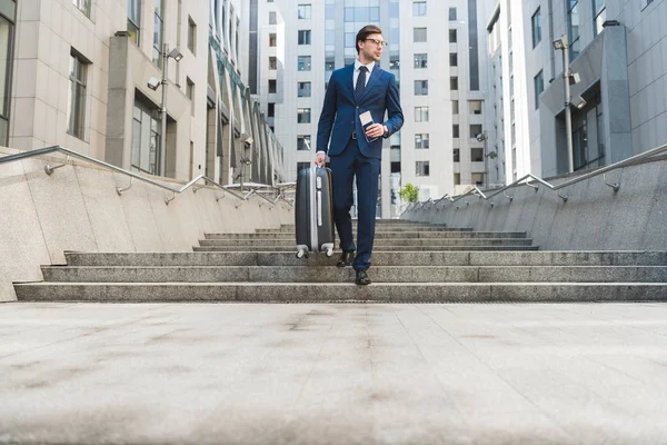 Attractive young businessman in stylish suit with luggage and flight tickets going down stairs in business district — Stock Photo